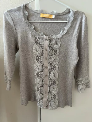 Grå kofta med spets från BIBA - Köpt på sellpy! Supersöt grå kofta från BIBA med trekvartsärmar och detaljerad spets längs knäppningen och halsringningen. Koftan har ribbad struktur och knappar framtill samt spetsdetaljer vid ärmsluten. Perfekt för en romantisk och feminin look.