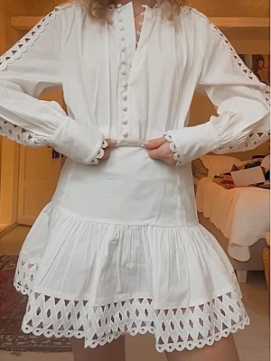 Studentklänning  - Superfin vit kortklänning med långärmade ärmar och dekorativa knappar framtill. Klänningen har spetsdetaljer längs ärmar och nederkant samt en volangad kjol. Perfekt för student - aldrig använd 
