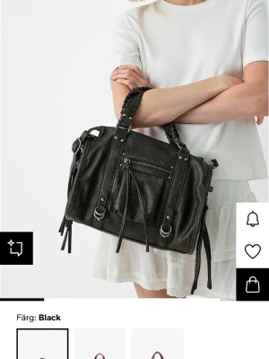 Svart handväska  - Cool svart handväska i konstläder med flätat handtag och flera dekorativa dragkedjor och remmar. Väskan har en rektangulär form och detaljer i silverfärg. 