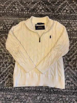 Vit kabelstickad tröja Polo Ralph Lauren - Kabelstickad vit tröja från Polo Ralph Lauren med halv dragkedja och hög krage. Tröjan har ribbade muddar och nederkant samt den klassiska broderade loggan på bröstet. Perfekt för dig som gillar stilrena och tidlösa plagg. Storlek M men passar även folk i S. Om ni har några frågor eller funderingar är det bara att höra av er! 