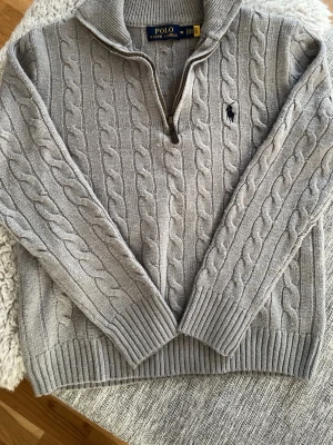 Polo Ralph Lauren - Kabelstickad grå tröja från Polo Ralph Lauren med halv dragkedja och hög krage. Tröjan har ribbade muddar och nederkant samt den klassiska broderade loggan på bröstet. Perfekt för lager på lager och riktigt snygg till jeans.