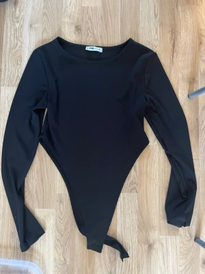 Svart långärmad ribbad body - Snygg svart body från NA-KD med lång ärm och rund halsringning. Bodyn är ribbad och har en klassisk, tight passform som framhäver figuren. Perfekt att styla med jeans eller kjol för en trendig look.
