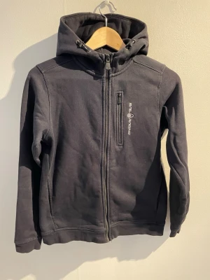 Svart hoodie från Sail Racing - Svart hoodie från Sail Racing med dragkedja framtill och en bröstficka med logga. Tillverkad i mjukt bomullsmaterial och har en stor huva med tryckt logotyp bak. Perfekt för en avslappnad och sportig stil.