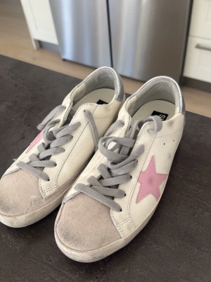 Golden Goose sneakers med rosa stjärna - Snygga vita Golden Goose sneakers med grå skosnören, grå mockadetaljer på tån och silverfärgad häl. På sidan finns en rosa stjärna som ger skon en unik touch. Tillverkade i läder och mocka, perfekta för en trendig streetstyle-look. Skorna är inte äkta men ser identiska ut, säljer pga de var för små för mig. Använd 1 gång 