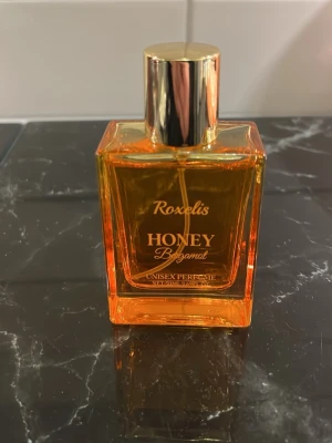 Roxelis Honey Bergamot Unisex Parfym - Fräsch unisex parfym från Roxelis med doft av honung och bergamott. Flaskan är fyrkantig i orange glas med en snygg guldfärgad kork. Perfekt för dig som gillar söta och citrusaktiga dofter.