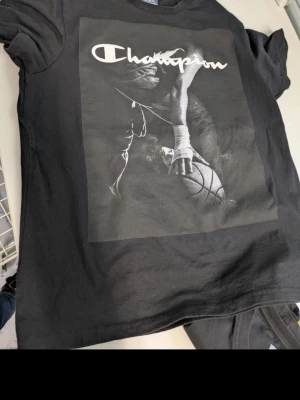 Svart Champion t-shirt med baskettryck - Cool svart t-shirt från Champion med stort tryck på framsidan som visar en basketspelare och loggan i vitt. Klassisk passform och rund hals. Perfekt för dig som gillar sportig streetstyle.