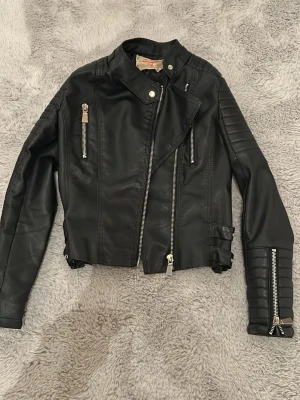 Svart biker skinnjacka - Snygg svart skinnjacka i bikerstil med asymmetrisk dragkedja, quiltade detaljer på ärmarna och flera silverfärgade dragkedjor framtill. Jackan har ståkrage med tryckknapp och justerbara spännen i midjan + lite puffigare axlar. Jackan är från märket Voyelles och är några år, men väldigt fint skick, de enda jag lagt märke till är defekten på sista bilden, annars väldigt bra skick. :))