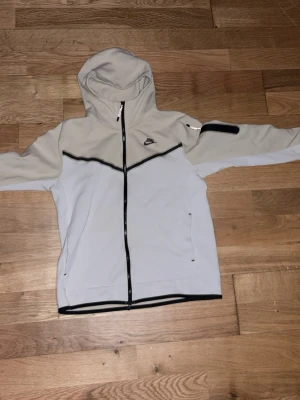 Nike hoodie i beige och vitt med dragkedja - Nike hoodie i beige och vitt med svarta detaljer och hel dragkedja framtill. Hoodien har en sportig look med huva och svarta linjer som accent. Tillverkad i mjukt material, perfekt för en chill och avslappnad stil. Inte så använd mest hängt i garderoben eftersom jag köpte fel storlek hör gärna av er om frågar:)