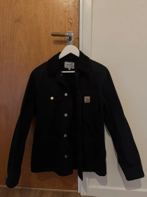 Svart jacka från Carhartt - Svart jacka från Carhartt med klassisk krage, raka ärmar och knäppning framtill. Jackan har en bröstficka med Carhartt-logga och guldiga knappar som snygga detaljer. Materialet är slitstarkt och jackan har en enkel, tidlös design.