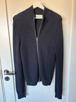 Mörkgrå stickad ziptröja från Casual Friday - Snygg mörkgrå stickad tröja från Casual Friday med dragkedja hela vägen framtill. Tröjan har hög krage, ribbstickade muddar och är tillverkad i ett grovt stickat material. Perfekt för lager-på-lager och stilren look.