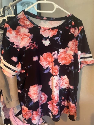 Blommig topp med korta ärmar - Säljer en svart topp med stora blommor i orange och rosa nyanser. Toppen har korta ärmar med randiga detaljer och en rund halsringning. Perfekt för en färgglad look!