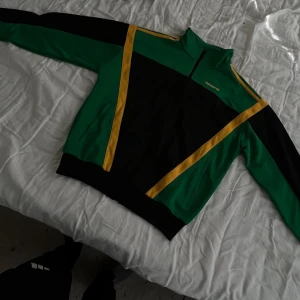 Grön och svart track jacket från Adidas - Säljer en retroinspirerad track jacket från Adidas i grönt och svart med gula detaljer. Dragkedja framtill, ståkrage och två fickor. Perfekt för dig som gillar sportig streetwear och vill sticka ut med färg.