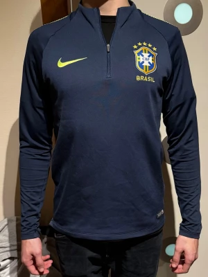 NIKE BRAZIL TRÄNINGSTRÖJA - Mörkblå långärmad träningströja från Nike med Brasilien CBF-logga och gul Nike-swoosh på bröstet. Halvlång dragkedja framtill och gula detaljer på axlarna. Perfekt för fotbollsträning eller sportiga aktiviteter.                                                       (Modellen är 180cm, 72kg)