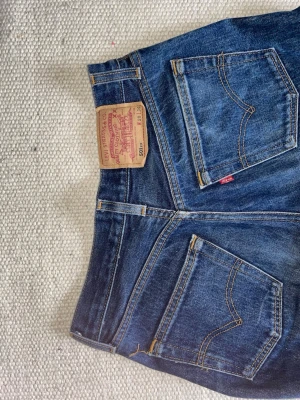 Levi's 501 raka blå jeans - Klassiska Levi's 501 jeans i mörkblå denim med raka ben och femficksmodell. Jeansen har den ikoniska läderlappen bak i midjan och röda Levi's-taggen på bakfickan. Tillverkade i bomull med snygga kontrastsömmar och knappgylf.