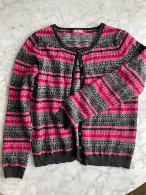 Mjuk mönstrad kofta - Mjuk o skön kofta från Indiska med rund hals och knäppning framtill. Långa ärmar och ribbade muddar. Koftan är rosa, cerise, vit o grå i ullblandning och med gulligt mönster.