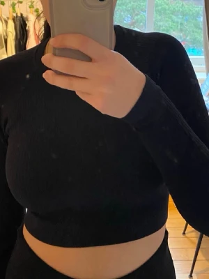 Svart croppad långärmad topp - Säljer en svart croppad topp med lång ärm och rund hals. Toppen är ribbad och har en tight passform som framhäver figuren. Perfekt till höga jeans eller kjol. Materialet känns mjukt och stretchigt, troligen bomull eller polyester.