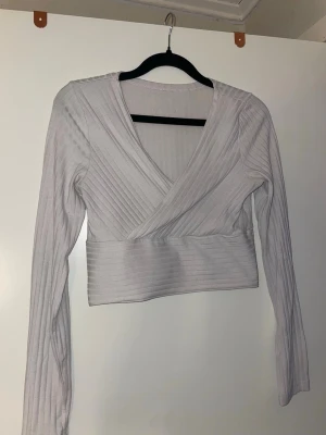 Ljusribbad omlottopp med v-ringning - Trendig ljus omlottopp med v-ringning och ribbad struktur. Toppen har långa ärmar och en croppad passform som ger en modern look. Perfekt att styla med höga jeans eller kjol.