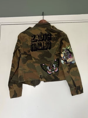 Jeansjacka från Zadig & Voltaire - Cool camouflagemönstrad jacka från Zadig & Voltaire med broderade detaljer och patchar, bland annat fjäril och texten 'Malibu'. Jackan har lång ärm, dragkedja och flera fickor framtill. 