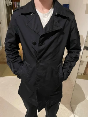 Svart trenchcoat från Stone Island - Snygg svart trenchcoat från Stone Island med klassisk dubbelknäppning och axelklaffar. Jackan har två sidofickor och det ikoniska Stone Island-märket på vänster ärm. Perfekt för dig som gillar stilren och tidlös design. Nypris:13,300
