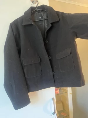 Svart overshirt från LIMITD - Stilren svart overshirt från LIMITD med klassisk krage och fyra knappar framtill. Två stora fickor med lock på bröstet ger en cool utility-look. Overshirten är gjord i ett mjukt, tjockare tyg som passar perfekt till lager-på-lager-stil.