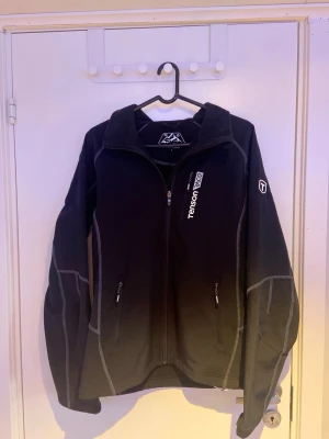 Svart vindjacka från Tenson - Snygg svart vindjacka från Tenson med vit logga på bröstet och ärmen. Jackan har dragkedja framtill, två fickor med dragkedja och grå kontrastsömmar. Tillverkad i ett lätt och funktionellt material, perfekt för blåsiga dagar.