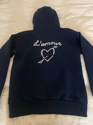 Mörkblå hoodie med L'amour-tryck från ginatricot  - Mörkblå hoodie med huva och vitt tryck på bröstet och ryggen där det står 'L'amour' tillsammans med ett hjärta och pil. Säljer då den inte passar mig❤️Orginalpris är ca 279kr skriv för mer bilder eller frågor.🥰använd ett fåtal gånger.(på bilden är snörena i luvan borttagna men kommer så klart sättas tillbaka vid köp)💕pris kan diskuteras!💕