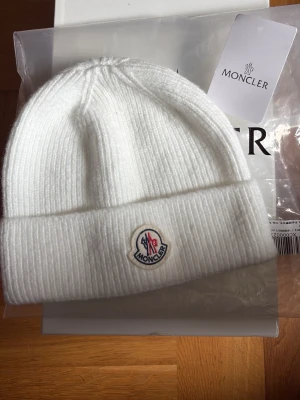 Moncler mössa - Snygg vit ribbstickad mössa från Moncler med uppvikt kant och klassisk Moncler-logga framtill. Tillverkad i mjukt material som håller dig varm och stilren under kalla dagar. Perfekt accessoar för dig som vill ha en clean och exklusiv look.