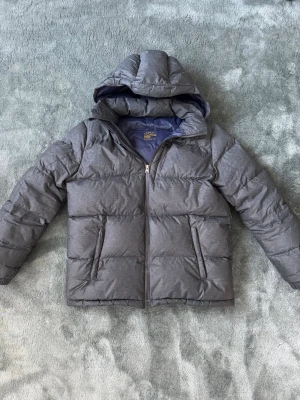 Grå Ralph lauren puffejacka - Snygg grå pufferjacka från Lindex med vadderad design och stor huva. Jackan har dragkedja framtill, två sidofickor med dragkedja och quiltade sömmar. Perfekt för kalla vinterdagar och ger en modern look.