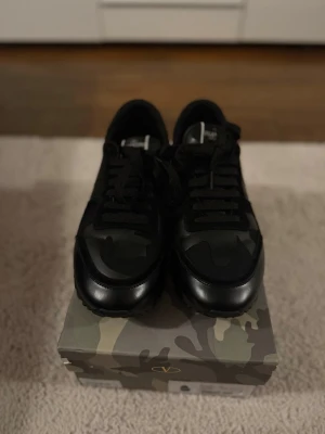 Svarta Valentino Garavani sneakers - Snygga svarta sneakers från Valentino Garavani med kamouflagemönster på tån och detaljer i läder och textil. Skorna har svarta skosnören och en klassisk låg siluett. Perfekta för dig som vill ha en stilren men ändå unik look.