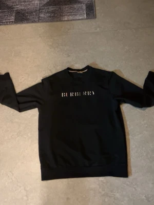 Svart Burberry hoodie med logga - Snygg svart hoodie från Burberry med klassisk logotyp tryckt på bröstet. Tröjan har rund halsringning och ribbade muddar vid ärmslut och nederkant. Perfekt för en stilren och trendig look.