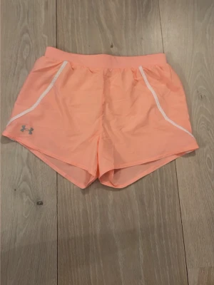 Persikofärgade träningsshorts Under Armour - Säljer ett par persikofärgade träningsshorts från Under Armour med vita detaljer och logga på vänster ben. Shortsen har elastisk midja med dragsko och är tillverkade i ett lätt och snabbtorkande material, perfekt för intensiva träningspass.