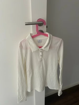 Vit ribbad långärmad henley - Säljer en vit ribbad henley med lång ärm och krage. Skjortan har tre knappar framtill och är gjord i ett mjukt, stretchigt material som känns bekvämt. Perfekt för dig som gillar en clean och enkel stil.