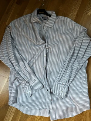 Randig skjorta från Tommy Hilfiger - Snygg vit och blå randig skjorta från Tommy Hilfiger med klassisk krage och långa ärmar. Skjortan har knappar framtill och vid ärmsluten samt diskret logga på manschetten. Modellen är fitted och passar perfekt till en stilren look. Stl 45/18 ca en L eller XL 