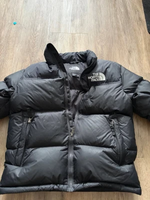 Svart pufferjacka från The North Face - Snygg svart pufferjacka från The North Face i storlek XL junior, vilket motsvarar 152-160 cm enligt deras storlekstabell. Jackan har två fickor på sidorna och stor logga på bröstet och ryggen. Passa på att köpa en vanligtvis dyr jacka till ett lägre pris på grund av en knappt synlig lagning framtill (se bild 4).
