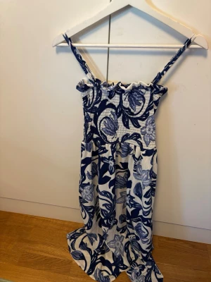 Blåvit blommig sommarklänning - Supersöt blåvit klänning med blommigt mönster och smock upptill. Klänningen har smala axelband och är perfekt för varma dagar. Lätt och luftig modell som ger en avslappnad känsla. Passar perfekt till sommaren!