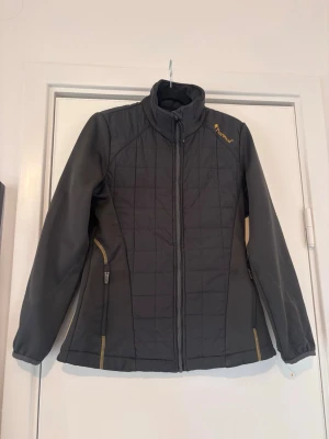 Svart vindjacka från Pinewood - Säljer en svart hybridjacka från det populära märket Pinewood. En mycket praktisk jacka som kombinerar vadderade partier på fram- och baksida för värme, med stretchiga ärmar och sidopaneler för optimal rörlighet. • Märke: Pinewood • Färg: Svart med snygga guld/orange kontrastsömmar. • Egenskaper: Två dragkedjeförsedda sidofickor, hög krage och figurnära passform. Skick: Jackan är i väldigt bra skick förutom en skada vid ena ärmslutet, som inte syns om man viker lite, eller om man syr ihop