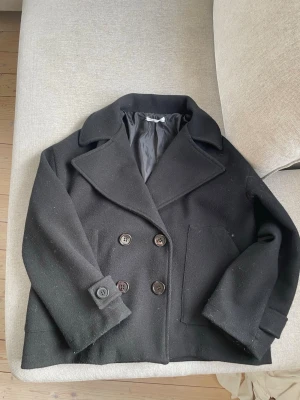 Svart kappa, Designby Si - Klassisk svart kappa med dubbelknäppning och stora slag. Jackan har två framfickor, dekorativa knappar vid ärmsluten och är fodrad. Perfekt för dig som gillar tidlös och stilig look. Endast varsamt använd och i mycket bra skick! 🤗💕