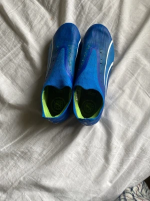 Blåa Puma fotbollsskor utan snörning - Säljer ett par blåa fotbollsskor från Puma med strumpliknande passform. Skorna har vita detaljer på sidorna och neonfärgade inslag på dobbarna och insidan. Ovandelen är i syntetmaterial och sulan är anpassad för spel på gräs. Köptes för 900 kr 