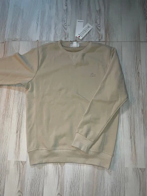 Beige sweatshirt från Lacoste - Stilren beige sweatshirt från Lacoste med rund halsringning och diskret broderad krokodil-logga på bröstet. Tröjan har ribbade muddar vid ärmslut och nederkant. Perfekt för en avslappnad och trendig look.