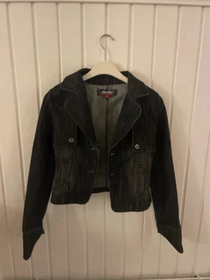 Svart jeansjacka från Onado - Snygg svart jeansjacka från Onado med markerad krage och två fickor framtill med knappar. Jackan har en croppad passform och coola sömdetaljer som ger en edgy vibe. Perfekt för dig som vill ha en klassisk men ändå unik jeansjacka i garderoben.