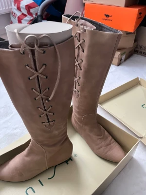 Beiga knähöga snörstövlar från Unisa - Snygga knähöga stövlar från Unisa i beige med spetsig tå och snörning framtill. Stövlarna har en stilren design och är perfekta för att lyfta din outfit. Materialet känns mjukt och exklusivt.