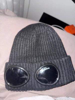 Svart C.P. Company goggle-mössa - Säljer en svart ribbstickad mössa från C.P. Company med ikoniska goggle-linser framtill. Mössan är i mjukt stickat material och har en uppvikt kant där linserna sitter. Perfekt för dig som vill sticka ut med en streetwear-inspirerad look.