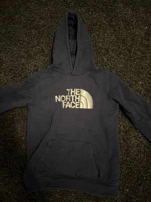 Mörkblå hoodie The North Face - Mörkblå hoodie från The North Face med stor vit logga på bröstet. Klassisk känguruficka framtill och ribbade muddar vid ärmslut och nederkant. Tillverkad i mjukt bomullsmaterial och har en stor huva för extra komfort.