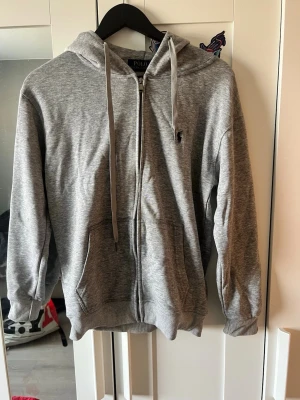 Grå zip hoodie från Polo Ralph Lauren - Snygg grå hoodie med dragkedja från Polo Ralph Lauren. Klassisk design med huva, dragsnören och fickor framtill. Liten broderad logga på bröstet. Tillverkad i mjukt bomullsmaterial som är skönt att ha på sig.