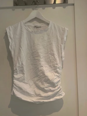 Vit ärmlös topp från Zara - Säljer en enkel och stilren vit topp från Zara. Modellen är ärmlös med rund hals och har en lätt rynkad detalj på sidorna. Perfekt för dig som gillar en clean look och vill ha något som är lätt att matcha. Materialet är mjuk bomull.