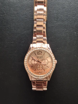 Roséguld klocka - Stilren klocka från Regal i roséguld med länkarmband och glittrande stenar runt urtavlan. Urtavlan har tre dekorativa små ur och stora siffran 12. Perfekt accessoar för att lyfta din outfit. Ståll frågor om man har.