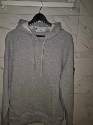 Grå hoodie från Strand Island - En stilren grå hoodie från Strand Island med klassisk huva och dragsnören. Hoodien har en känguruficka framtill och ribbade muddar vid ärmslut och nederkant. Perfekt för en avslappnad look. Materialet känns mjukt och bekvämt. Passar S bäst 