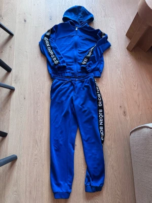 Blåa mjukisbyxor och huvtröja från Björn Borg - Träningsoverall från Björn Borg med svarta och vita logoremsor längs sidorna. Perfekta för chill eller träning, riktigt sköna och snygga. Stl 36.