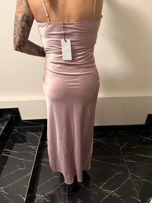 Ljusrosa långklänning från Sisters Point balklänning - Snygg ljusrosa långklänning från Sisters Point i glansigt material. Klänningen har smala axelband, omlott framtill och figurnära passform. Perfekt för dig som vill sticka ut med en elegant och trendig look.