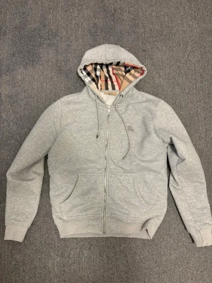Grå hoodie med dragkedja från Burberry - Snygg grå hoodie med dragkedja från Burberry. Klassisk huva med det ikoniska rutmönstret på insidan, dragsko och två fickor framtill. Tillverkad i mjukt bomullsmaterial, perfekt för en avslappnad och trendig stil.